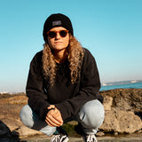 Black Beanie from It's okay | made in Portugal with love | feel cozy and warm with this knitted beanie. Pt gorro de inverno it's okay em preto. Image: women in squat position near the beach wearing black waffle beanie.