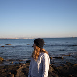 Blue Beanie from It's okay | design in Portugal with love | feel cozy and warm with this knitted beanie | gorro feito em Portugal, wear it with pride. Image: woman looking left wearing blue waffle beanie, sea behind her