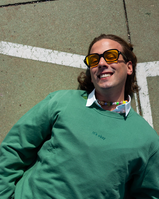 it's okay crewneck: green made in portugal with organic cotton. Join us. Image: boy on the floor wearing sunglasses and a necklace smiling