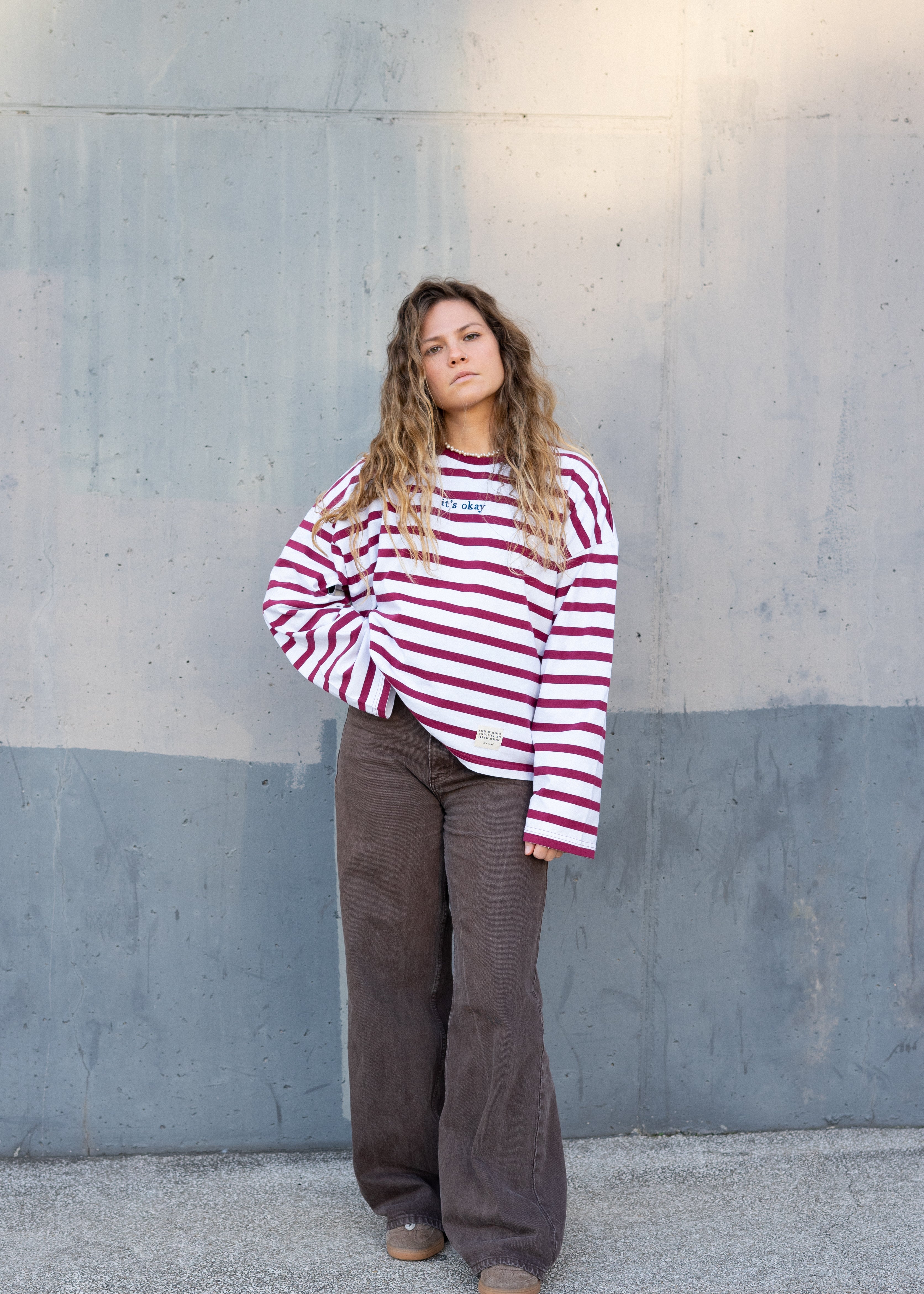 Girl wearing a red and white striped shirt and brown pants standing against a gray wall.