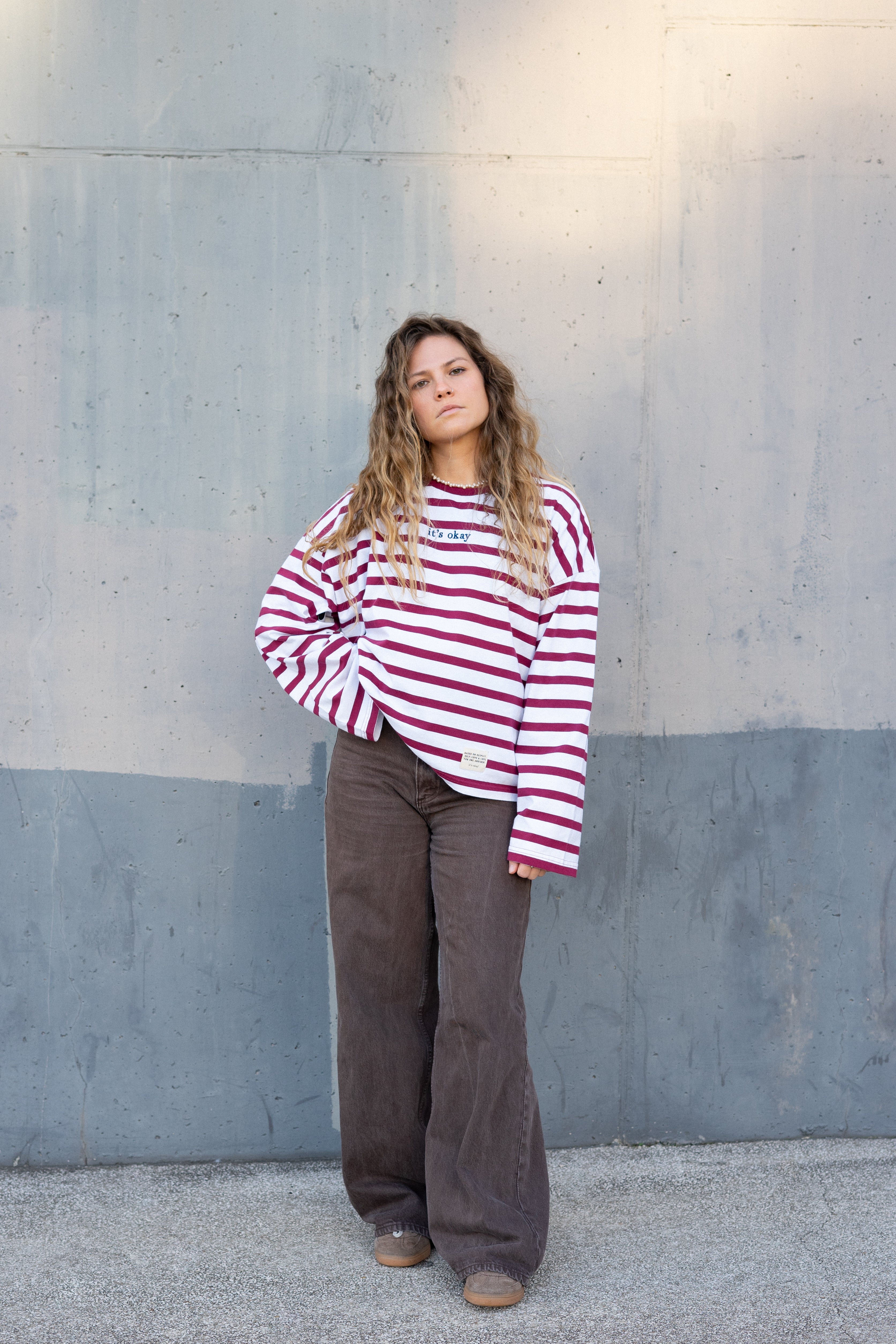 Girl wearing a red and white striped shirt and brown pants standing against a gray wall.