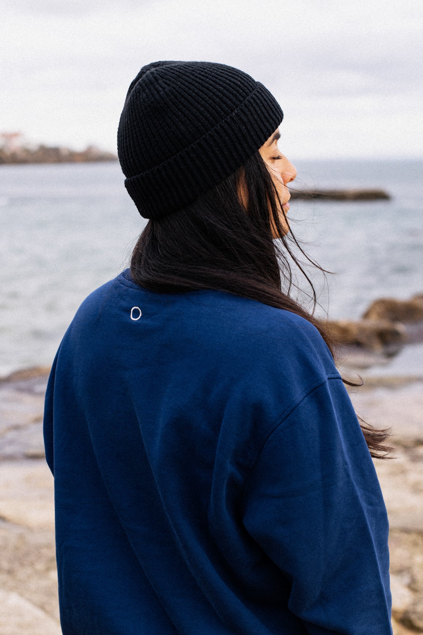 Back view of woman wearing navy IT’S OKAY organic cotton crewneck with embroidered circle, outdoors by the sea.