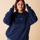 A person wearing a blue crewneck sweatshirt with embroidery on the front and back, posing with one hand raised to their head.
