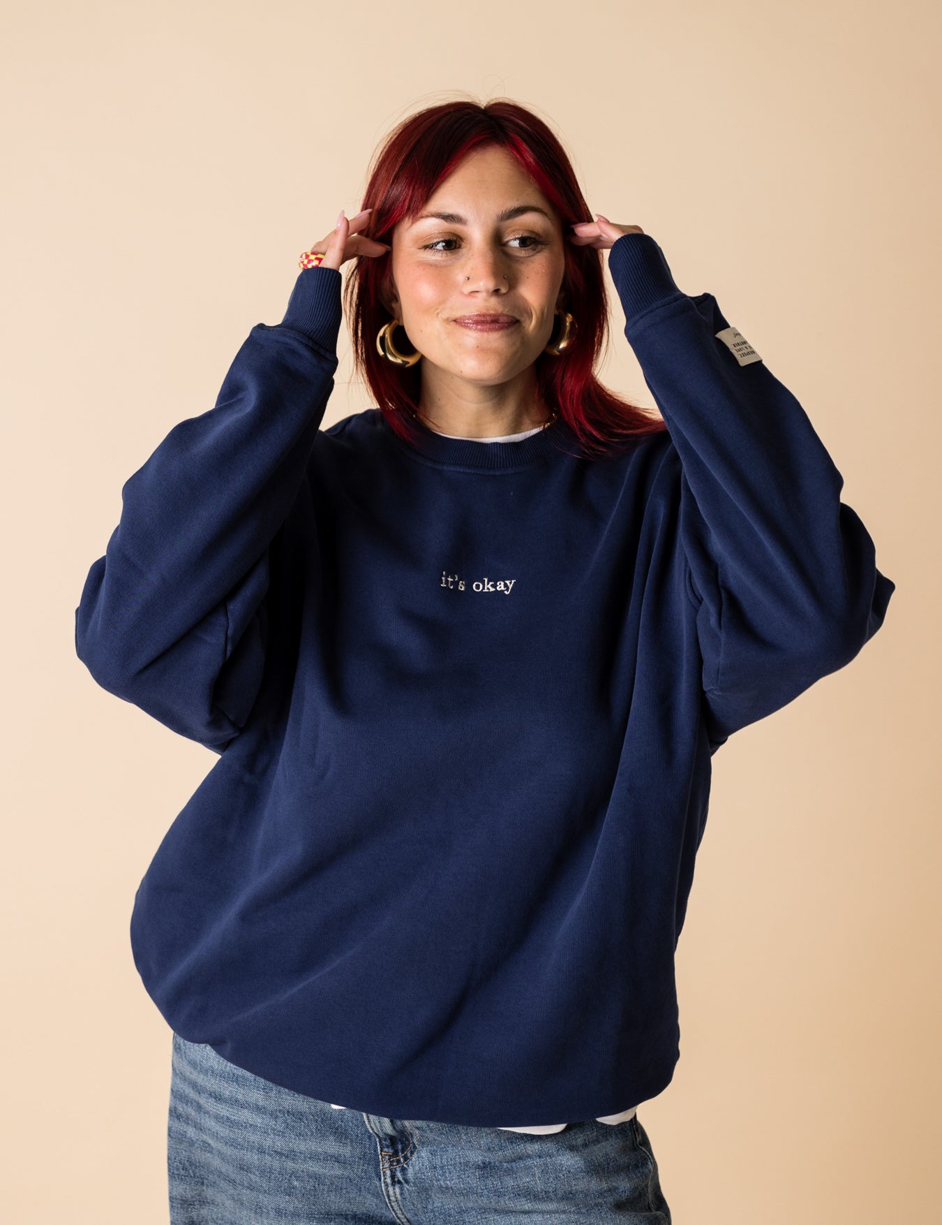 A person wearing a blue crewneck sweatshirt with embroidery on the front and back, posing with one hand raised to their head.