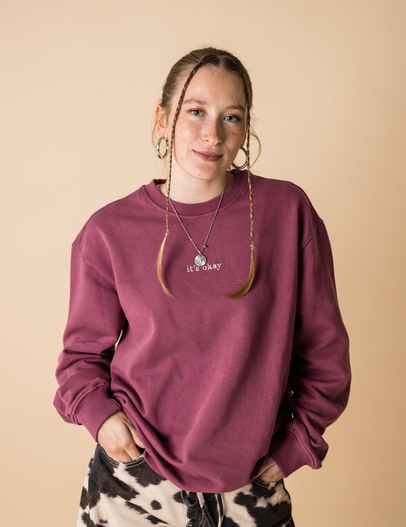 A person wearing a purple crewneck sweatshirt with a front and back embroidery, standing against a beige background.
