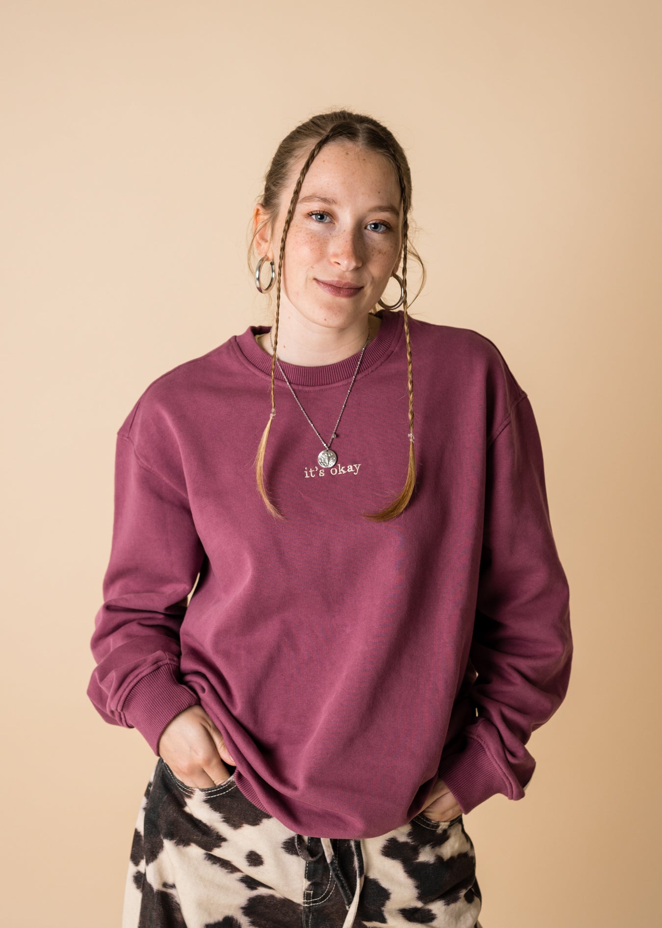 A person wearing a purple crewneck sweatshirt with a front and back embroidery, standing against a beige background.