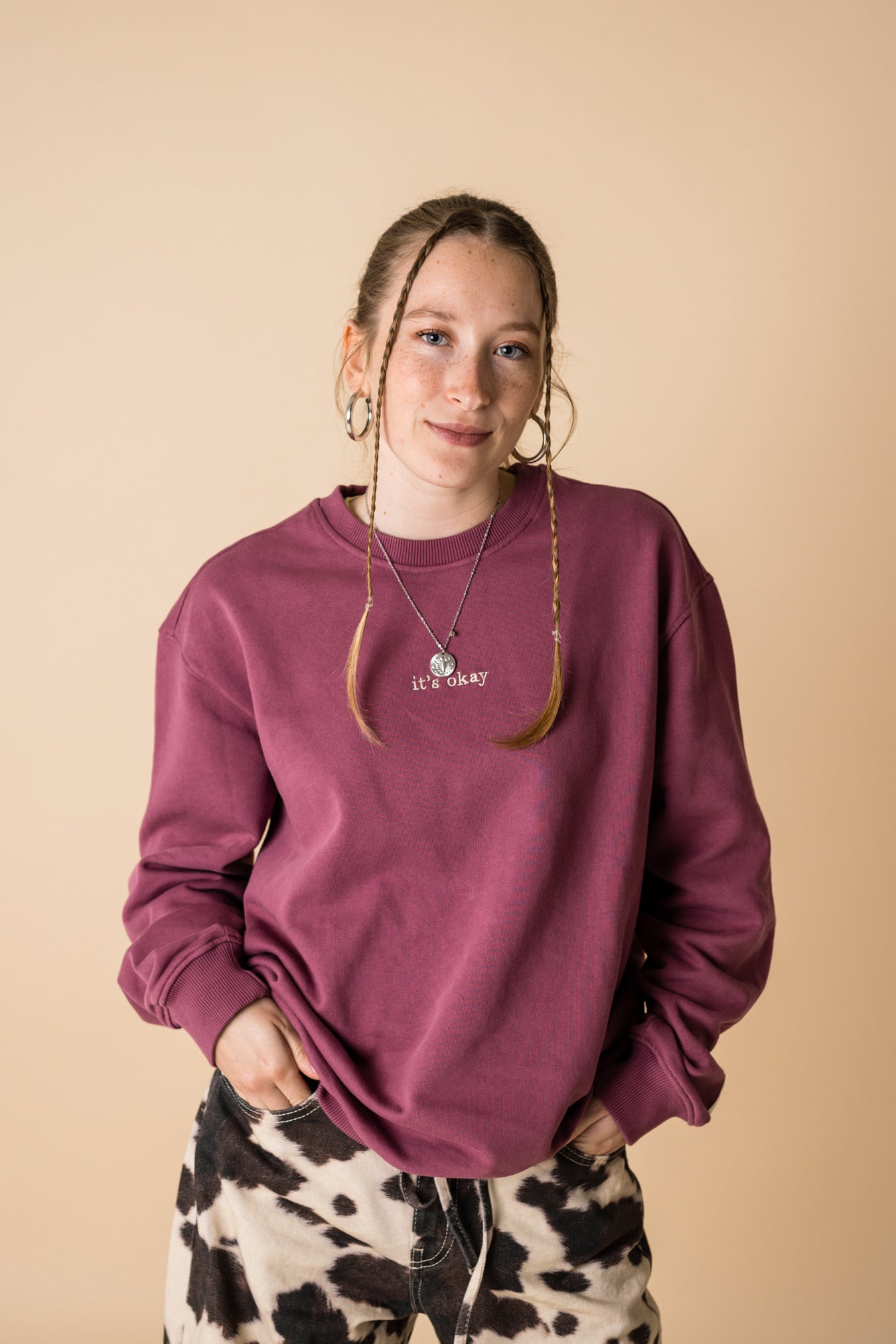 A person wearing a purple crewneck sweatshirt with a front and back embroidery, standing against a beige background.