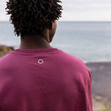 Back view of model wearing burgundy IT’S OKAY organic cotton crewneck with embroidered circle, outdoors by the sea.