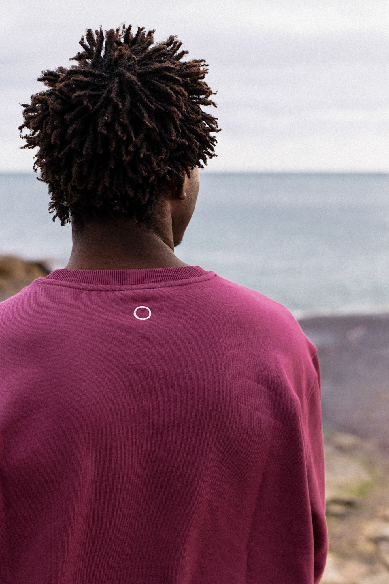 Back view of model wearing burgundy IT’S OKAY organic cotton crewneck with embroidered circle, outdoors by the sea.