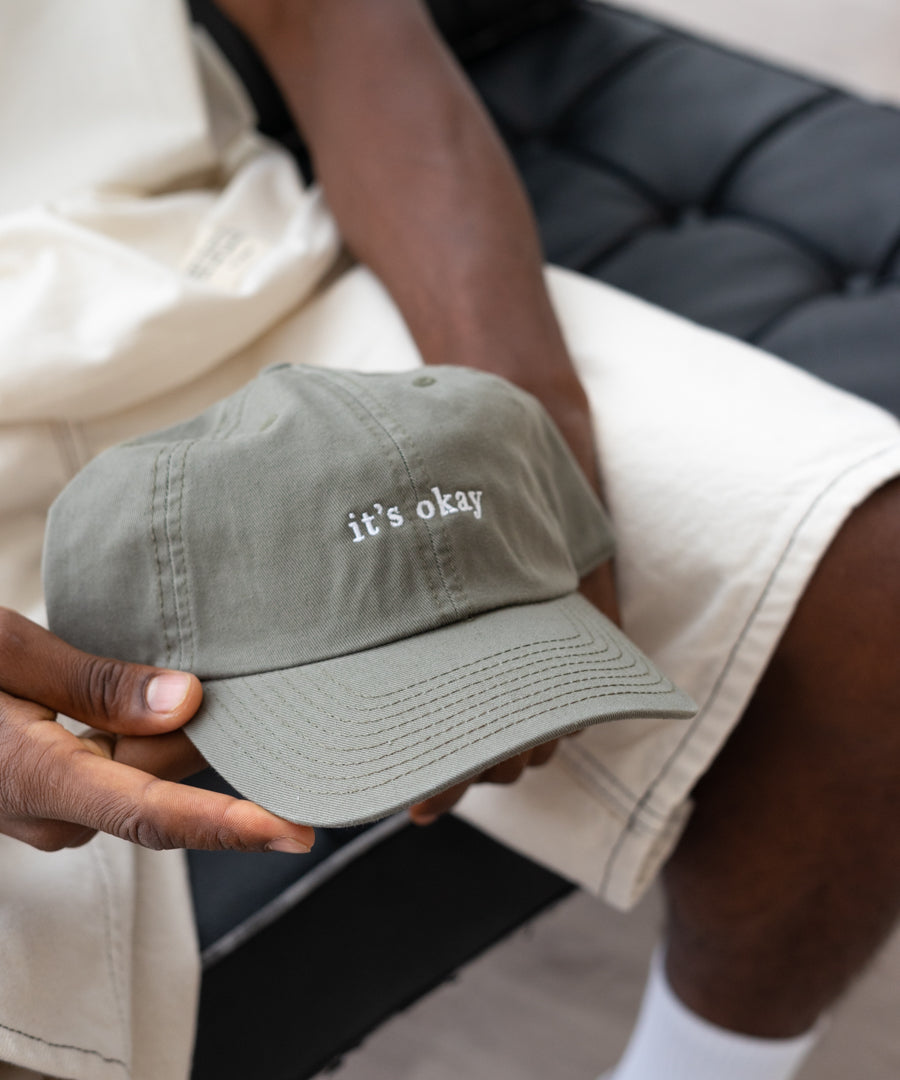 it's okay organic cap green | made with organic cotton, embroidered. The hat is on a black men's hands, he is wearing white shorts. 