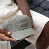 it's okay organic cap green | made with organic cotton, embroidered. The hat is on a black men's hands, he is wearing white shorts. 