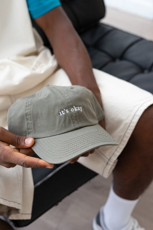 it's okay organic cap green | made with organic cotton, embroidered. The hat is on a black men's hands, he is wearing white shorts. 