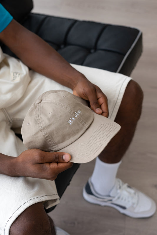 it's okay organic cap khaki | made with organic cotton, embroidered. The hat is on a black men's hands, he is wearing white shorts. 