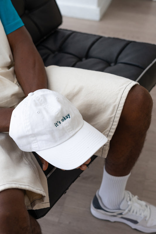 it's okay organic cap white | made with organic cotton, embroidered. The hat is on a black men's hands, he is wearing white shorts. 