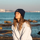 Blue Beanie from It's okay | design in Portugal with love | feel cozy and warm with this knitted beanie | gorro feito em Portugal, wear it with pride. Image: woman wearing blue waffle beanie with the sea behind her. 