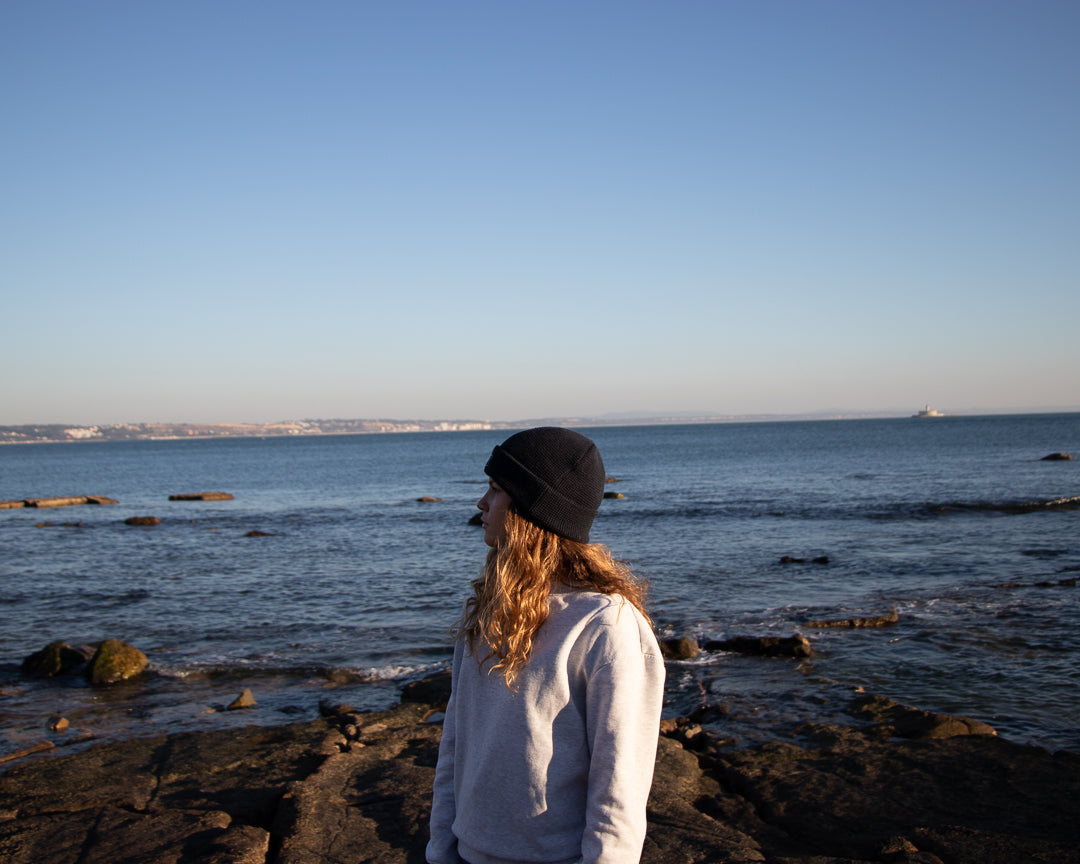 Blue Beanie from It's okay | design in Portugal with love | feel cozy and warm with this knitted beanie | gorro feito em Portugal, wear it with pride. Image: woman looking left wearing blue waffle beanie, sea behind her 