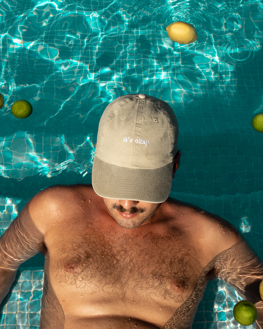 it's okay stone cap, beige cap made with 100% cotton, embroidered. it's okay to be exactly who you are. From Portugal with love. Boné de pala bege de algodão bordado em Portugal. Image: boy inside a swimming pool wearing the it's okay stone cap, with lemons floating on the water. 
