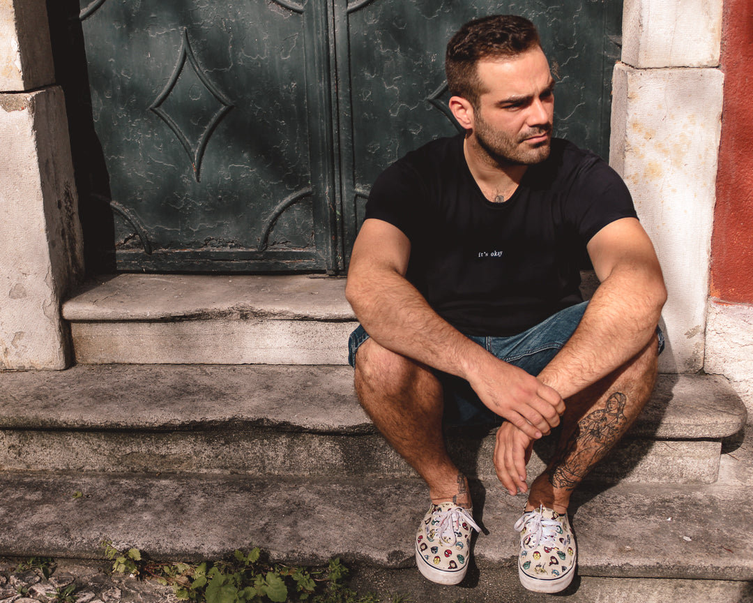 Organic t-shirt essential Black made in North of Portugal with love. Exclusive design made only for it's okay. T-shirt em algodão orgânico feito em portugal com amor | GOTS approved. Image: Guy sitting on a door stairs wearing the organic essential t-shirt 