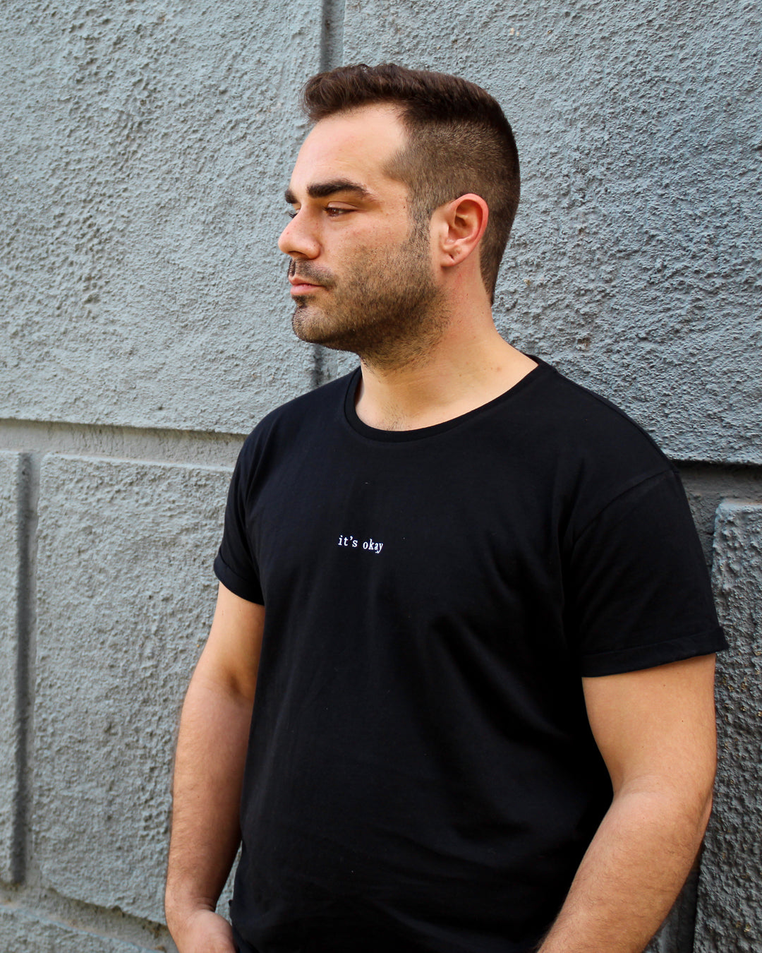 Organic t-shirt essential Black made in North of Portugal with love. Exclusive design made only for it's okay. T-shirt em algodão orgânico feito em portugal com amor | GOTS approved. Image: Guy standing up in a dark wall, wearing the essential black t-shirt made with organic cotton and love.