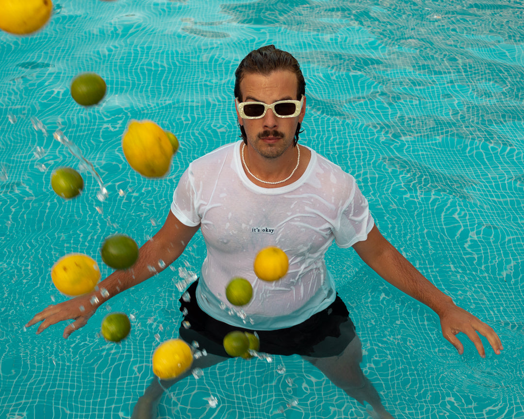 Organic t-shirt essential white made in North of Portugal with love. Exclusive design made only for it's okay. Pt. T-shirt em algodão orgânico feito em portugal com amor , design exclusivo| GOTS approved.I Image: Man inside  a swimming pool with white sunglasses and lemons being thrown 