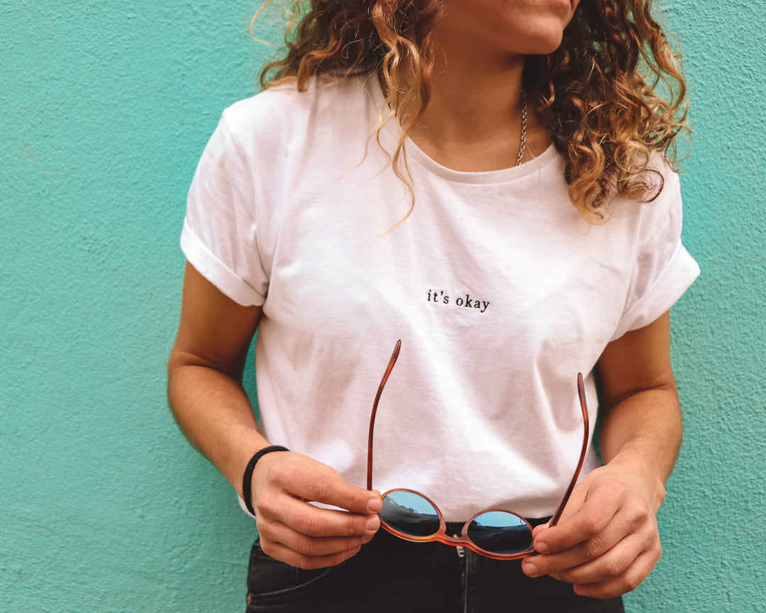 Organic t-shirt essential white made in North of Portugal with love. Exclusive design made only for it's okay. Pt. T-shirt em algodão orgânico feito em portugal com amor , design exclusivo| GOTS approved. Image: girl with sunglasses on her hand wearing the essential white t-shirt