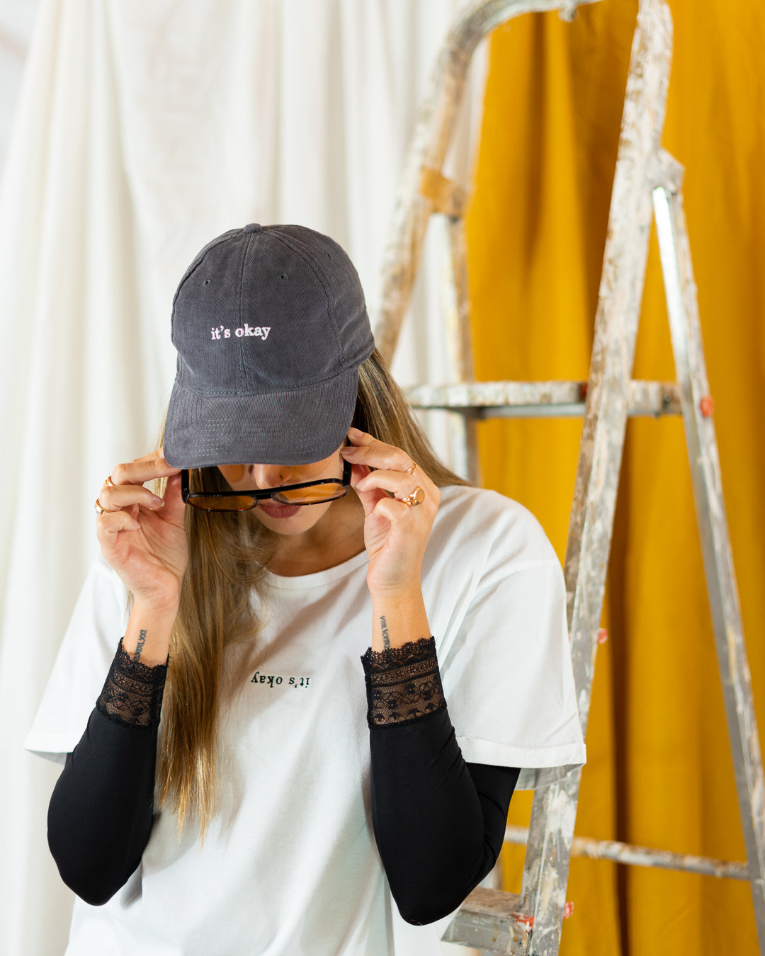 Storm cap | cotton needlecord corduroy in gray. Embroidered in Portugal. One size fits most | Wear it with pride. It's okay to be exactly who you are. Pt: Boné algodão de pala em bombazine de cor cinza, tamanha ajustável.  Image: girl with word search organic t-shirts from it's okay, taking of her sunglasses wearing a storm cap. In her back, a construction ladder.